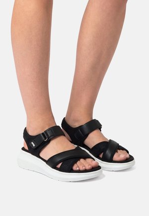 Piedi con sandali neri con punta aperta, cinturini regolabili e suole spesse bianche, in piedi su uno sfondo grigio chiaro.