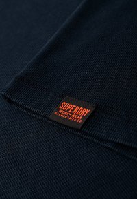 Tela texturizada en color marino que muestra un primer plano de una etiqueta tejida con "SUPERDRY" en naranja, unida al borde de una prenda.