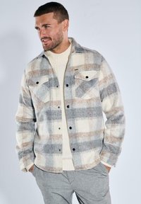 Karierter Fleece-Jacke in Grau, Creme und Beige, mit zwei Brusttaschen, schwarzen Knöpfen und einer weichen Textur, über einem cremefarbenen Strickpullover getragen.