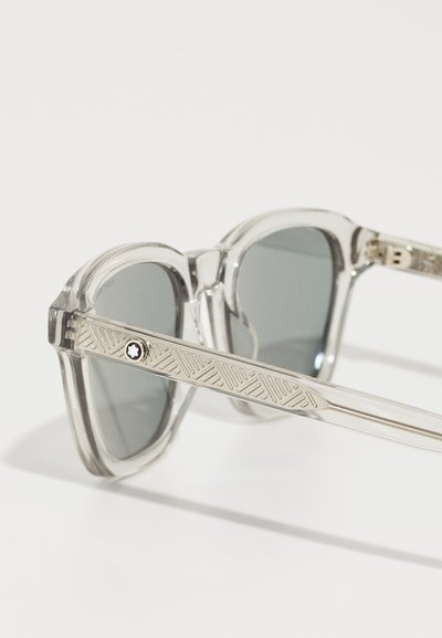 Lunettes de soleil en plastique transparent avec verres gris, dotées de branches en métal texturé et d'un emblème en forme d'étoile sur le côté, mettant en avant un design moderne.