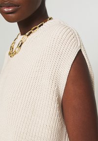 Béžový pletený svetr s texturovaným vzorem a otevřeným designem ramen, doplněný zlatým řetízkovým náhrdelníkem.