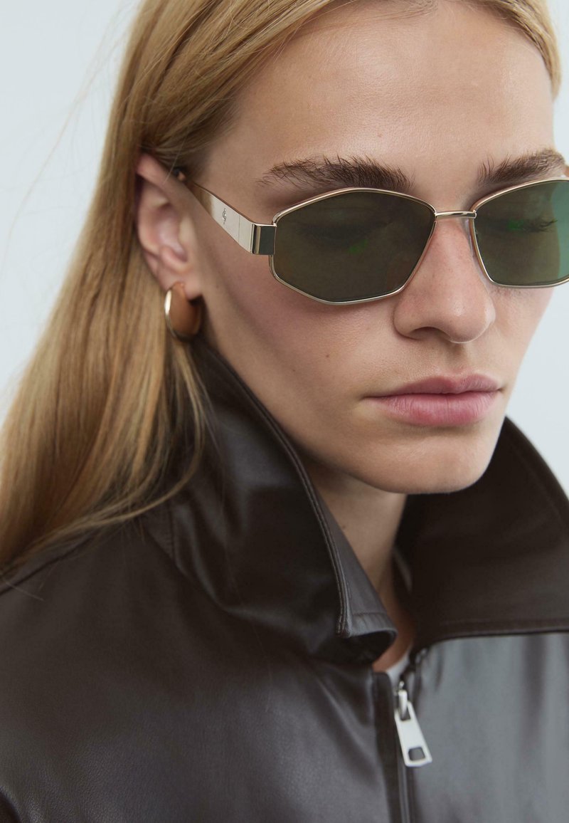 Sunglassar med oktagonala mörkgröna linser och metallbåge, med silverdetaljer. Modellen bär en svart läderjacka med hög krage.