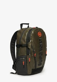 Sac à dos vert olive avec des accents noirs, deux poches latérales et des tirettes de zipper oranges. Présente un grand logo à l'avant et des bretelles rembourrées.