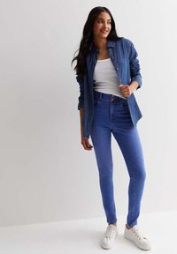 Dżinsowa koszula na białym topie na ramiączkach, zestawiona z niebieskimi dżinsami z wysokim stanem i białymi sneakersami. Gładki materiał, dopasowany fason, casualowy wygląd.