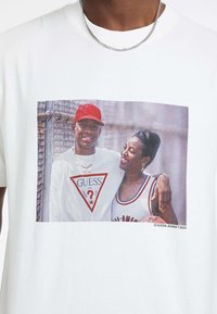 Biała bawełniana koszulka z graficznym nadrukiem na przodzie, przedstawiającym dwie osoby, czerwoną czapkę baseballową, trójkątny logo "GUESS" oraz piłkę do koszykówki.