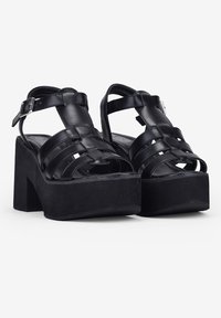 Sandalias de plataforma negras con un tacón grueso, que cuentan con correas de cuero trenzado, cierre con hebilla y una textura suave.