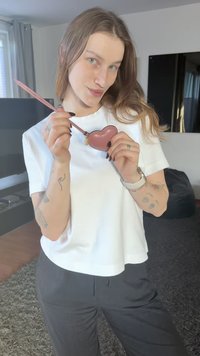 Accessoire rose en forme de cœur avec une fermeture éclair et un petit cadenas, tenu avec un crayon rose. Le porteur, vêtu d'un haut blanc et d'un pantalon noir, se tient à l'intérieur.
