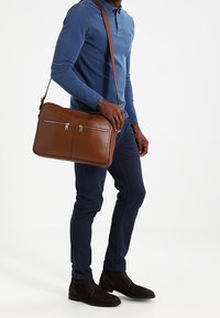 Mężczyzna w niebieskiej koszuli i spodniach, trzymający brązową skórzaną torbę na ramię z przednimi kieszeniami na zamek, stojący na białym tle.