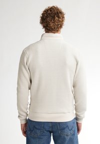 Svetle béžová mikina na zip s texturovanou látkou, žebrovaným límcem a manžetami a spodním lemem. Nosí se s modrými džíny, zobrazena zadní strana.