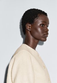 Profielweergave van een persoon met kort, krullend haar die een crèmekleurige, gestructureerde jas draagt tegen een effen lichte achtergrond.