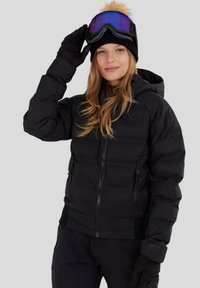 Svart pufferjacka med en figursydd design, dragkedjestängning och justerbar huva. Matchas med en svart mössa och purpurfärgade goggles.