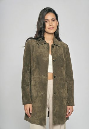 Kvinde med langt mørkt hår iført olivengrøn ruskindsjakke over lys crop top og beige bukser, stående foran en ensfarvet grå baggrund.