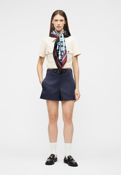 Γυναίκα που φοράει κρεμ κοντομάνικο cropped πουκάμισο, μεταξωτό φουλάρι με σχέδια, σκούρο μπλε σορτς με ψηλή μέση, λευκές κάλτσες και σκούρα δερμάτινα loafers.