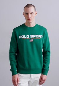 Sweatshirt en coton vert avec col rond, arborant le texte "POLO SPORT" en blanc et rouge ainsi qu'un graphique du drapeau américain. Manches longues, coupe décontractée.