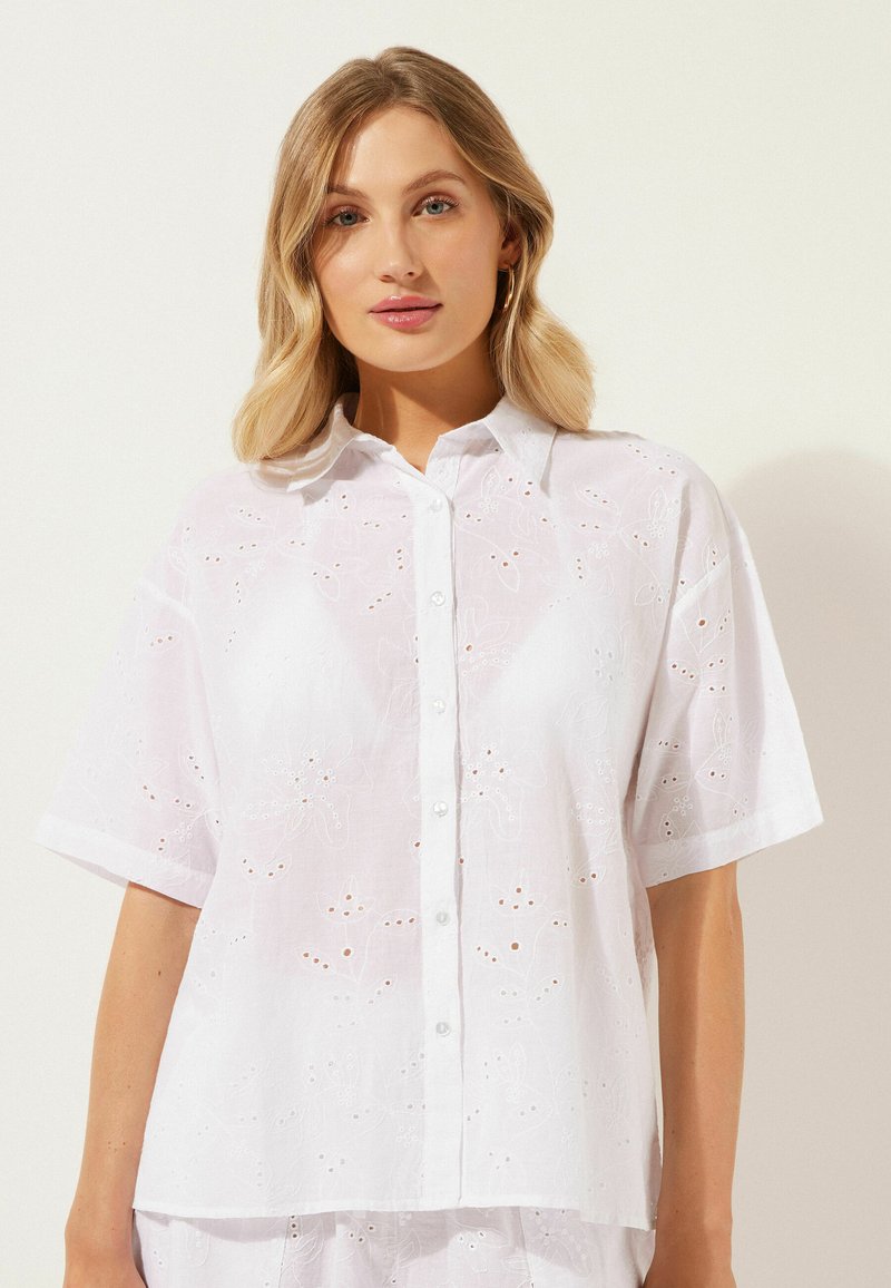 Camicia bianca a maniche corte con motivi floreali ricamati e tagli a occhio, con colletto e chiusura a bottoni.