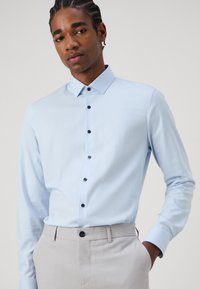 Cămașă albastră deschis cu mânecă lungă, cu guler cu butoni și accente închise la culoare, asociată cu pantaloni gri deschis. Textură netedă, design ajustat.