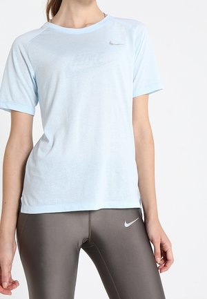 Kobieta ubrana w jasnoniebieską sportową koszulkę z krótkim rękawem i ciemnoszare legginsy Nike, stojąca z lewym ramieniem opuszczonym, a prawą ręką trzymająca brzeg koszulki.