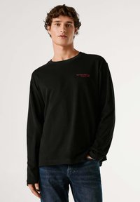 Črna majica z dolgimi rokavi in okroglim ovratnikom, z rdečim napisom na prsih. Iz mehke bombažne tkanine, kombinirana z modrimi jeans hlačami.