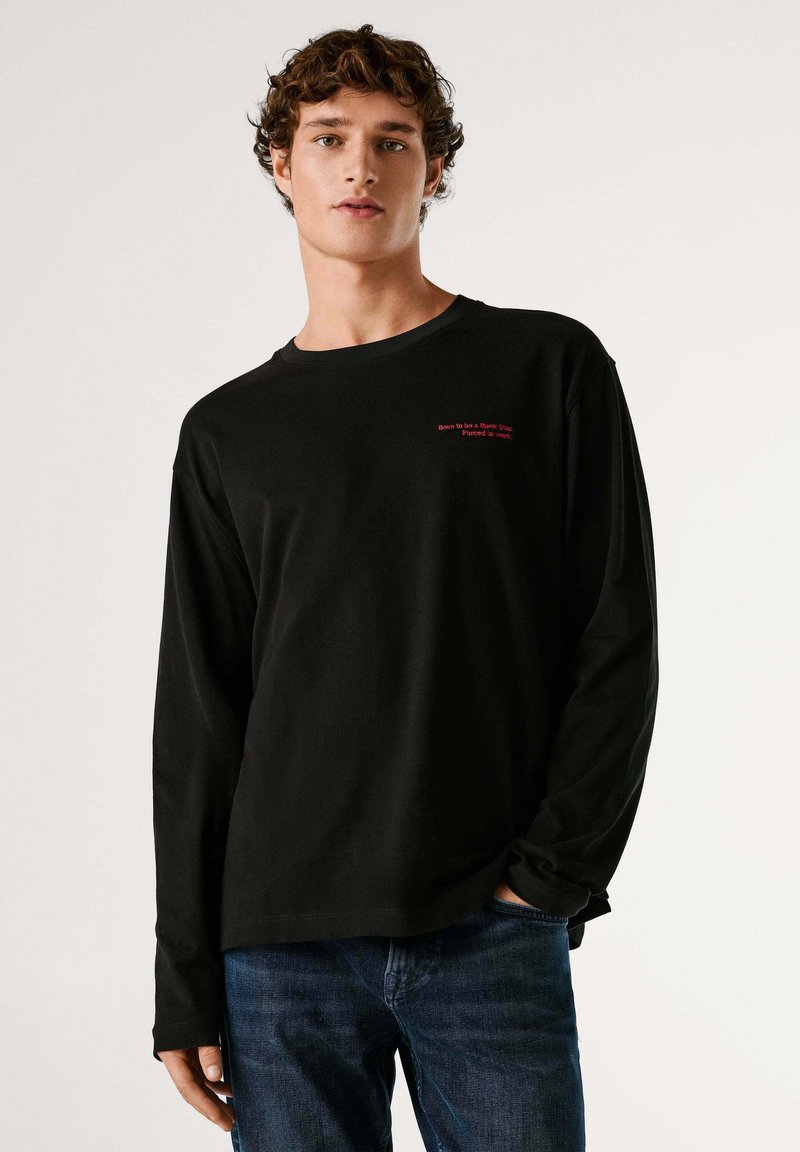 Črna majica z dolgimi rokavi in okroglim ovratnikom, z rdečim napisom na prsih. Iz mehke bombažne tkanine, kombinirana z modrimi jeans hlačami.