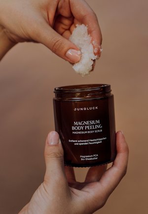 Braunes Glasgefäß mit der Aufschrift "Magnesium Körperpeeling", das in einer Hand gehalten wird, mit einer Handvoll weißem Peeling darüber. Glatte, glänzende Textur; minimalistisches Design.