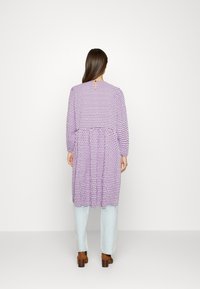 Vestido roxo de manga longa com bolinhas pretas, corte solto, cintura franzida, fecho nas costas em forma de chave, combinado com jeans azuis claros e saltos castanhos.
