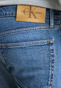 Plave traperice s smešnim kožnim oznakom označenom "Calvin Klein Jeans" s utisnutim logotipom, koje prikazuju klasični dizajn s pet džepova.