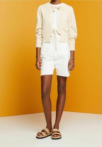 Personne portant un cardigan beige à manches longues avec des liens, un short blanc taille haute, des sandales beiges et des bracelets dorés, debout devant un fond jaune.