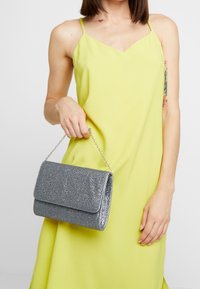 Silverglittrande clutchväska med lockstängning och kedjearmband, med rektangulär form och slät textur. Bärs tillsammans med en ljusgul klänning.