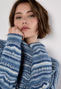 Young person with short brown hair wearing a blue patterned knit sweater, resting chin on hand against a plain white background.