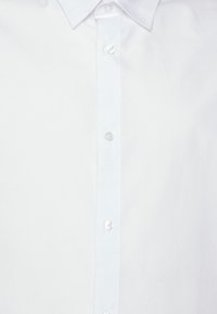 Camisa blanca de botonadura con cuello clásico. Tiene una textura suave y tejida, y cinco botones circulares y transparentes en la parte delantera.