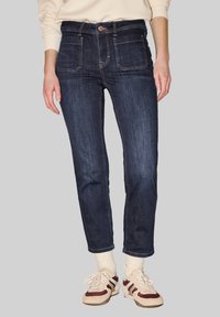 Jean slim bleu foncé porté avec des chaussettes crème et des baskets crème avec des rayures bordeaux et bleues, sur un fond gris.