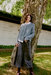 Cardigan grigio lavorato a maglia con grandi bottoni, camicia bianca con colletto e gonna in ecopelle scura. Stivali al ginocchio marroni completano l'outfit.