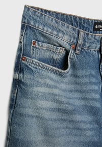 Jean en denim bleu clair avec une finition délavée, doté de cinq poches, de rivets en cuivre et de coutures orange le long des coutures.