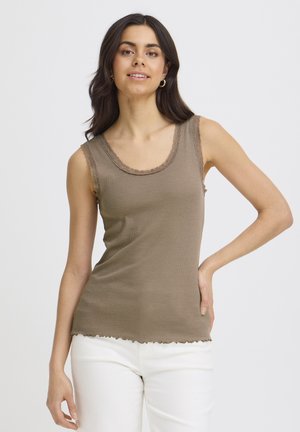 Junge Frau mit dunklem Haar, die ein ärmelloses braunes Oberteil mit Spitzenbesatz und weiße Hose trägt, posiert mit einer Hand in der Hüfte.