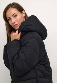 Melna pūkainā jaka ar kapuci, ar saplēstu dizainu un gludu tekstūru. Modelis pielāgo apkakli ar brīvu satvērienu.