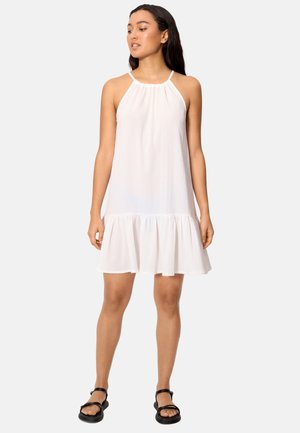 Mujer con vestido blanco sin mangas con cuello halter y dobladillo con volantes, combinado con sandalias negras abiertas, de pie frente a un fondo liso.