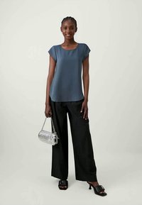 Blusa azul de manga curta, calças largas pretas e sapatos de salto abertos pretos. Bolsa de mão com corrente prateada e detalhes texturizados.