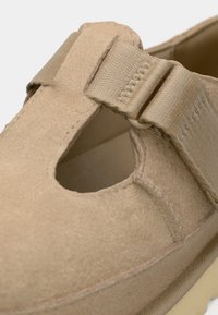 Beige suède schoen met een ronde vorm, voorzien van een uitsnijd ontwerp en een bandje voor sluiting. Gladde textuur en minimalistische esthetiek.