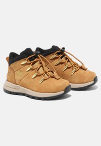 Chaussures de randonnée beige avec une tige en suède, un col noir, des détails en or et une semelle en caoutchouc. Elles disposent d'un système de laçage rapide et d'accents texturés.