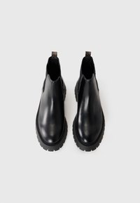 Une paire de bottes Chelsea en cuir noir avec des semelles épaisses et des languettes, présentée côte à côte sur un fond blanc.