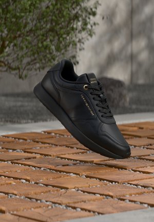 Sneaker nero Tommy Hilfiger con occhielli dorati e logo, che fluttua sopra un terreno in legno e con texture di ciottoli, con sfondo sfocato di piante.
