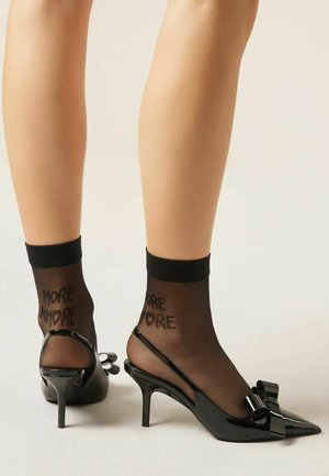 Gambe con calze nere trasparenti alla caviglia con la scritta "MORE AMORE" e décolleté slingback in pelle verniciata nera con dettagli a fiocco.