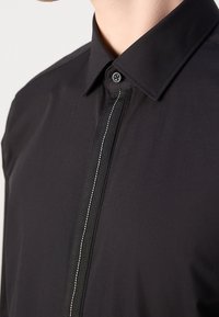 Camisa negra de vestir con cuello estándar, que presenta una raya vertical texturizada por el frente y un cierre de botones. Tejido suave.