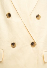 Μπεζ διπλό κουμπί σακάκι με τέσσερα κουμπιά χελώνας και γιακά με λοξή σχισμή, που δείχνει μέρος των εμπρόσθιων τσεπών.