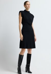 Schwarzes gestricktes Kleid mit kurzen, strukturierten Ärmeln und einer taillierten Silhouette. Kombiniert mit knielangen schwarzen Boots aus krokodilgeprägtem Leder.