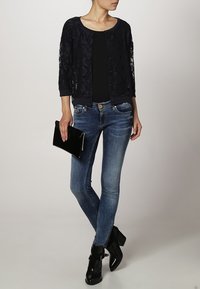 Cardigan in pizzo blu navy con maniche a tre quarti, top fitted nero, jeans skinny blu, pochette nera e stivali neri alla caviglia.