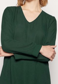 Grön V-ringad långärmad topp i mjukt tyg, med avslappnad passform, slät textur och viktade ärmar för extra komfort.