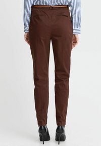 Braune Slim-Fit-Hosen mit glatter Textur, ausgestattet mit zwei Gesäßtaschen und einem Gürtel. Kombiniert mit schwarzen High-Heels und einem gestreiften Hemd.