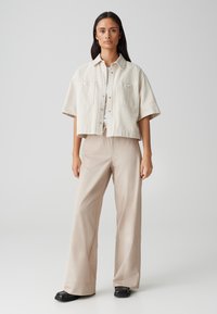 Veste cropped beige à manches courtes avec fermeture à boutons et deux poches poitrine, associée à un pantalon large beige clair et des chaussures noires.