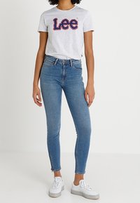 Camiseta de algodón gris con el logo "Lee" en rojo y azul, combinada con jeans ajustados en azul. Zapatillas blancas con suelas texturizadas.